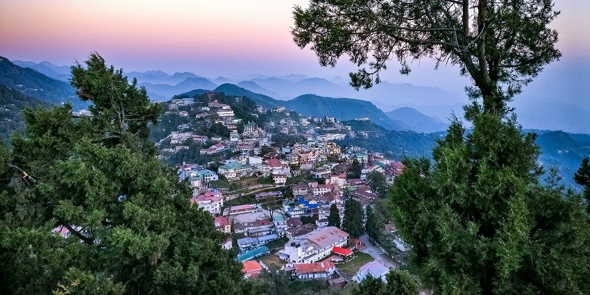 An image of a hilly town of Mussoorie, with buildings scattered across green slopes, surrounded by forests and mountains.