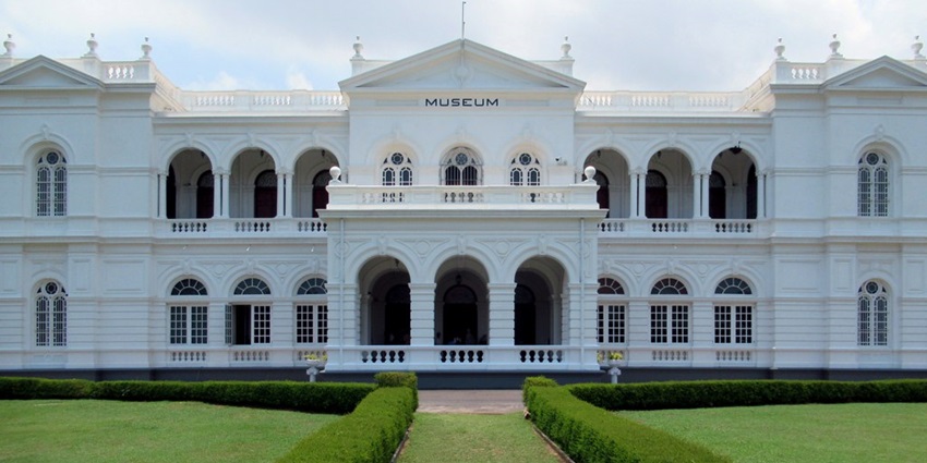 An image of the National Museum Colombo building with arches, columns, gardens, a paved path, and trees