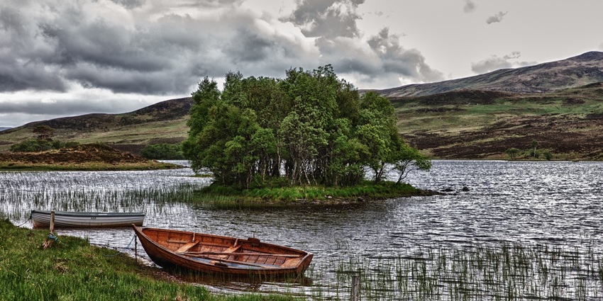 Peaceful lake with a small island, boats parked at the shore and hills in the background.