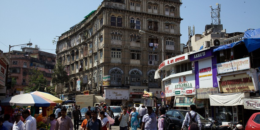 A bustling market street of Mumbai, filled with people, shops, and historic buildings.