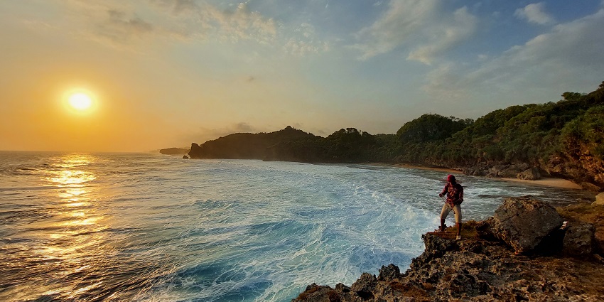 A person stands on rocky cliffs, watching a vibrant sunset over a scenic ocean bay.