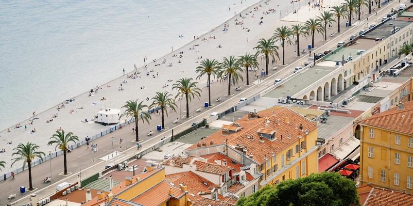 Promenade des Anglais in Nice with pebbled beach, blue sea, and colorful Mediterranean buildings.