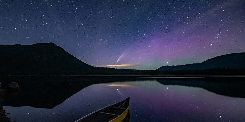 Yellow Canoe parked near the shore of a lake with a vibrant starry night sky and mountains