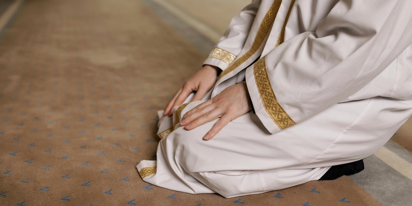 A person wearing a white robe with gold trim kneels on a prayer rug with a pattern