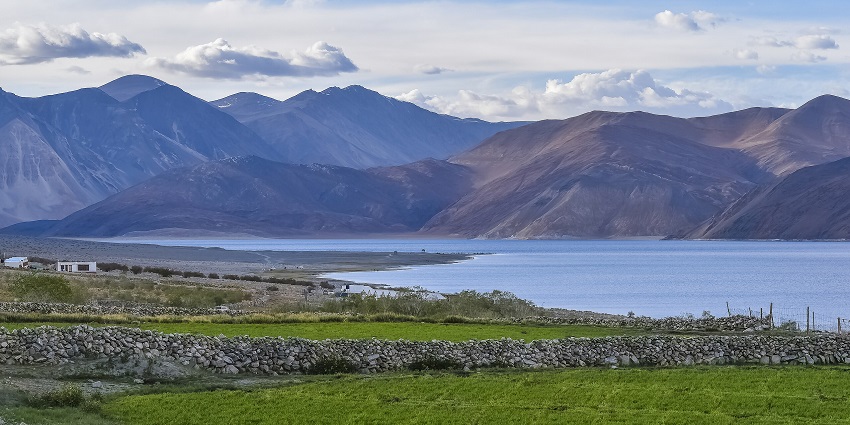 Pangong Tso lake with green plains, distant small buildings, sandy shorelines and mountains