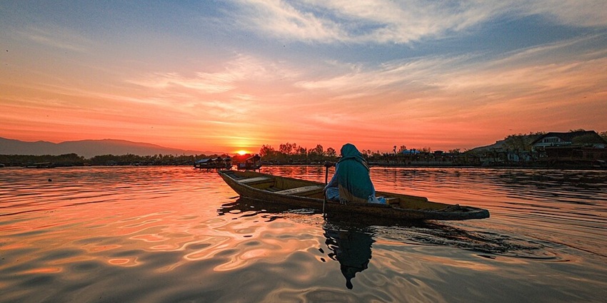 Dal lake with a large still water body, a woman on a wooden boat and a vibrant sunset