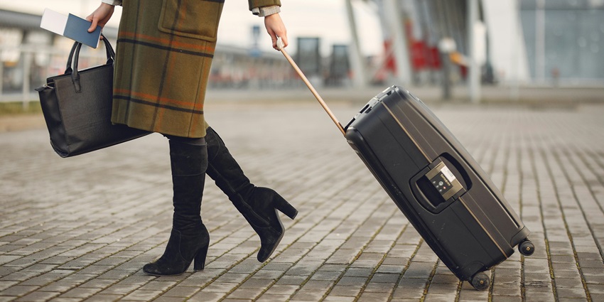 Traveller in a stylish coat and boots pulling a black suitcase outside an airport terminal