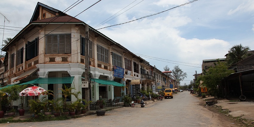 An image of a street in Kampot, Cambodia, with buildings, vehicles, and people in view.