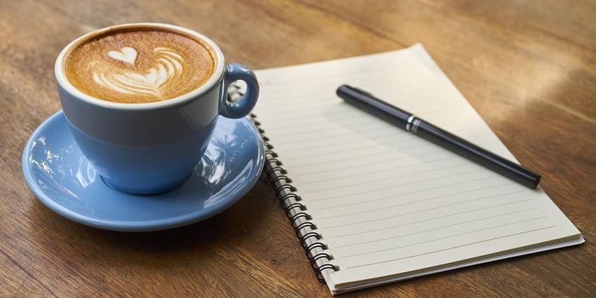 A view of a notebook kept on a brown table with a cup of coffee on the side.