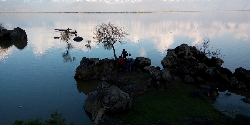 Wular lake with a rocky shoreline, people sitting on the rocks and in the distant boats