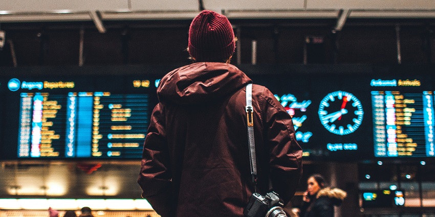 Image of a man standing infront of airport billboard for flights details - man hanging a camera