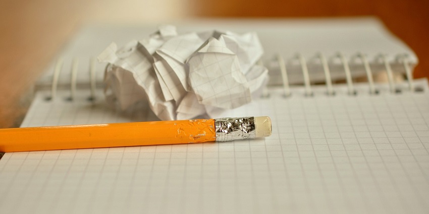A worn yellow pencil with eraser rests on a grid notebook beside a crumpled sheet of paper