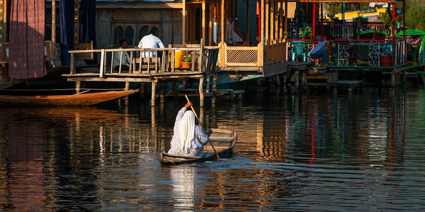 A woman boating in Dal Lake with clear water and people sitting outside the houseboats