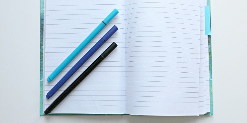 Open lined notebook with three pens in shades of blue and black resting on the pages.