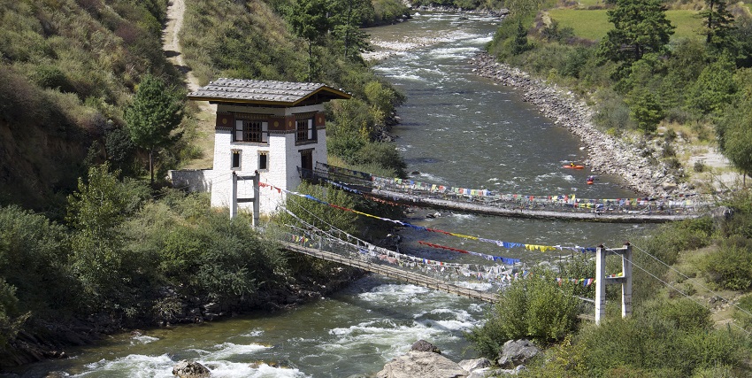 An image of kayaking in the Paro Chhu river in Bhutan, with two suspension bridges, prayer flags, and a white building