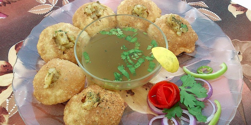 An image of a plate of Pani Puri, with six stuffed puris, a bowl of green liquid, and garnished vegetables