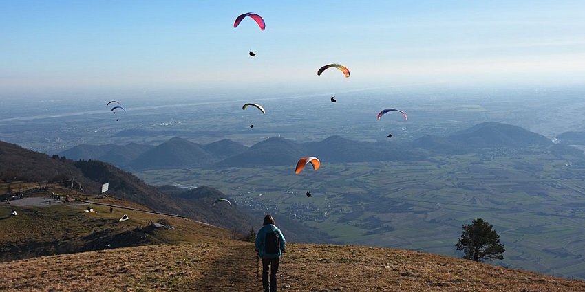 Person standing on a hill with base camp and multiple people paragliding mid-air