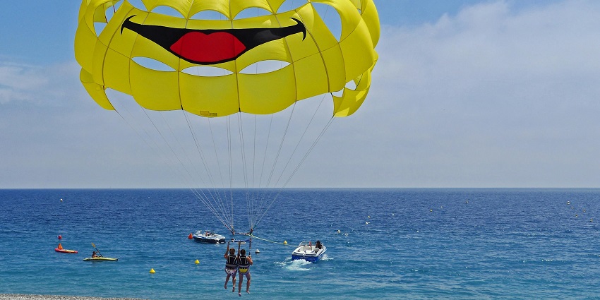 An image of two people harnessed below a bright yellow parasail with a smiley face.