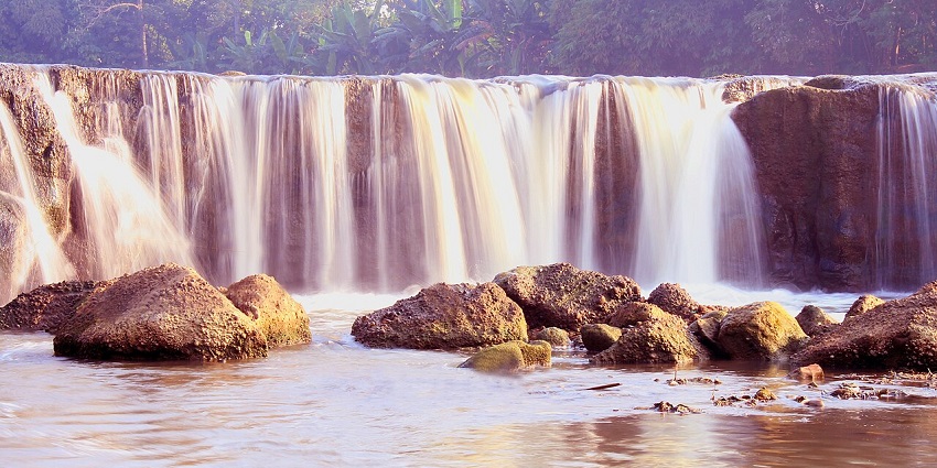 Water cascades over a wide rocky ledge at Curug Parigi waterfall, surrounded by greenery and natural riverbank scenery.