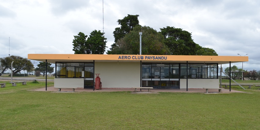 Paysandú Airport's small terminal is standing under a cloudy sky with trees behind it