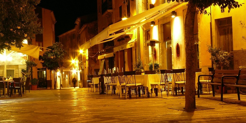 A cosy outdoor dining area is glowing under warm yellow lights on a quiet evening street.