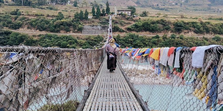 An image of a suspension bridge over the Pho Chhu river, with colourful prayer flags, rural terraced greenery, and buildings