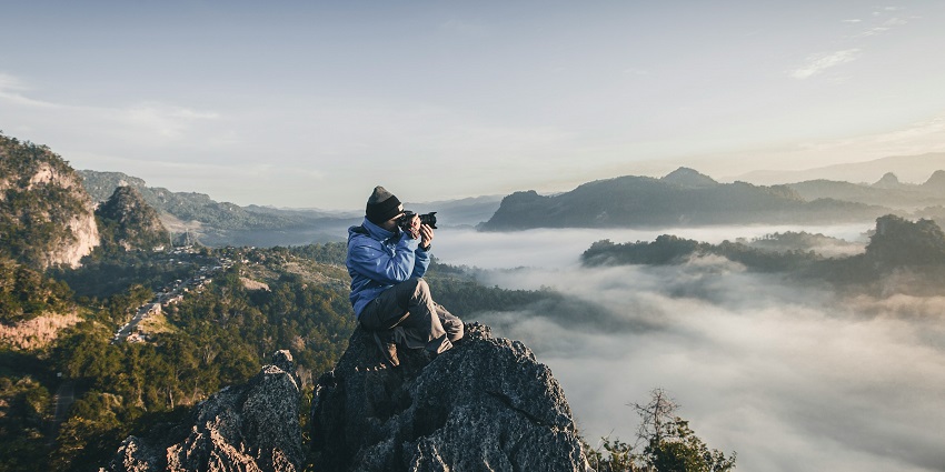 A photographer in a blue jacket sits on a rocky cliff, capturing misty mountains at sunrise.
