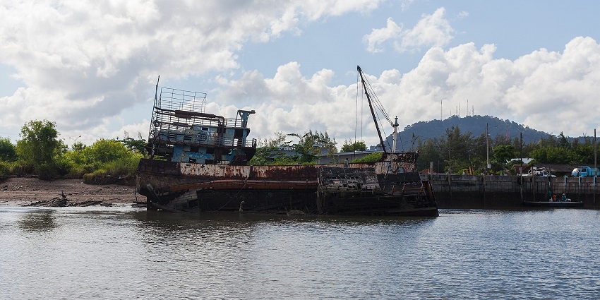 Rusting shipwreck partially submerged near the shore, surrounded by greenery and hills