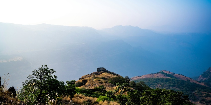 An image of a foggy, mountainous landscape with two hikers climbing one of the forts in the Rajmachi Trek, Maharashtra.