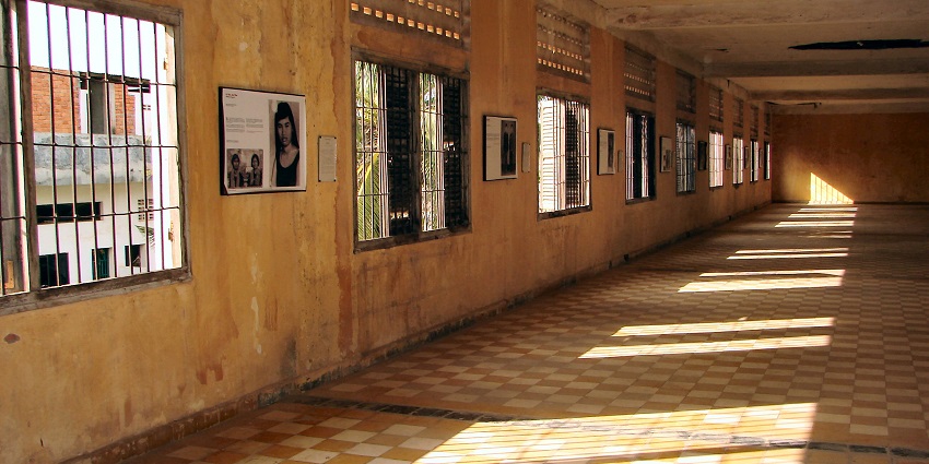 Image of a narrow corridor with barred windows, faded yellow walls, and checkered tiles illuminated by sunlight.