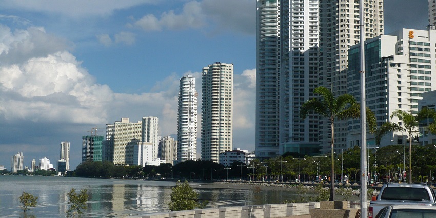 A waterfront cityscape with modern high-rise buildings and palm trees along the promenade
