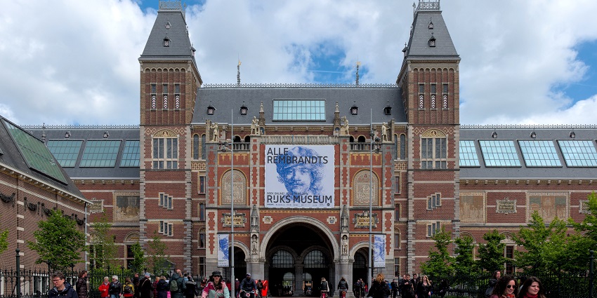 Daytime view of the Rijksmuseum in Amsterdam with its grand gothic-style facade, clear skies above, and people cycling through the central archway.
