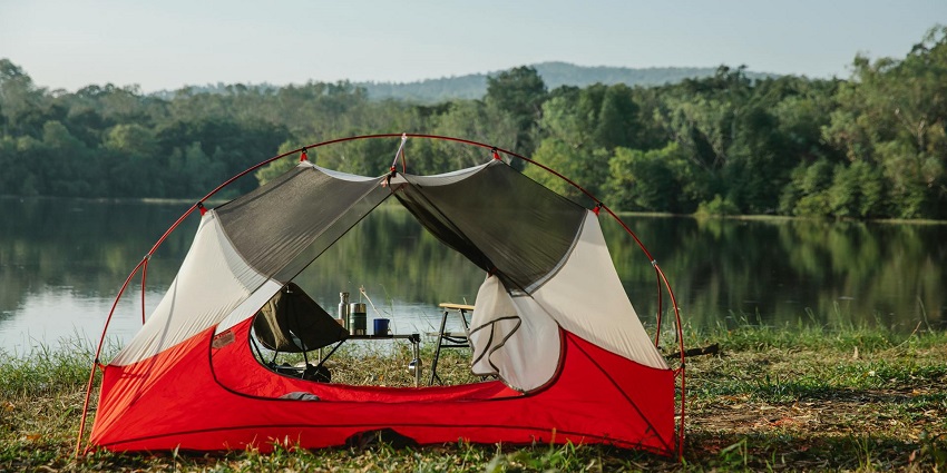 Red and white camp tent near the river with chairs, tables and green shoreline in the back