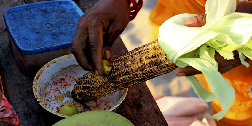 An image showing a roasted bhutta being seasoned by a seller with spices for added flavour.