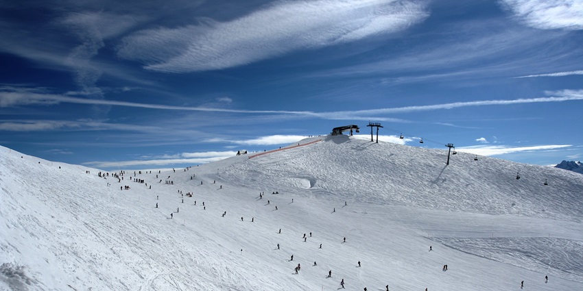 People ski as acres of snow sprawl across the mountain, and the blue sky with white clouds hangs above.