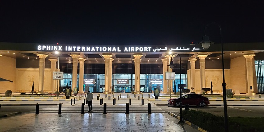 People are walking and cars are moving outside Sphinx International Airport at night.