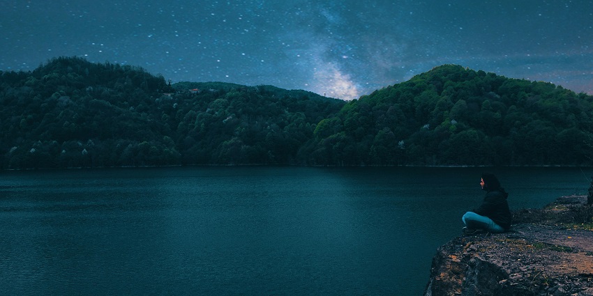 Woman sitting on a rocky cliff with bright stars in the sky and distant green mountains
