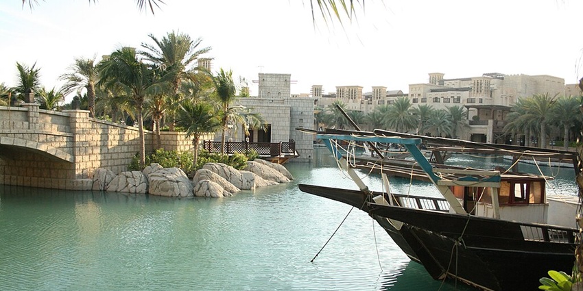 A Wooden boat docked on a turquoise canal with palm trees and stone bridge nearby.