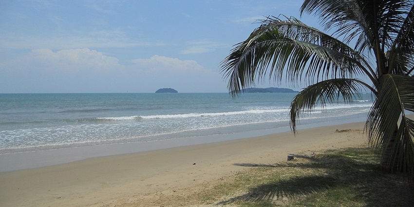 An image of the Tanjung Aru beach with a sandy shore, blue sky, gentle waves, palm trees, and distant islands
