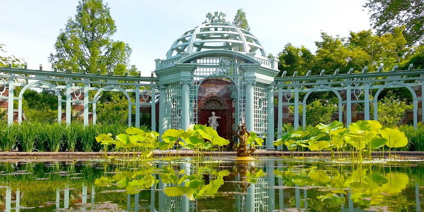A lush green garden around a lake with a white structure, a statue and a path for walking