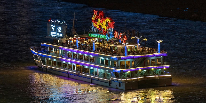A brightly lit river cruise boat at night, reflecting colourful lights on the water.
