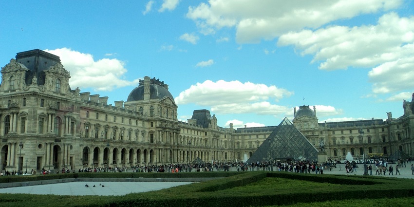 Front view of the Louvre Museum in Paris with its iconic glass pyramid and historic palace structure under a clear blue sky.