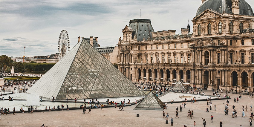 An image of the Louvre Museum in daylight, showcasing its ornate facade, statues, gardens, and cloudy skies.