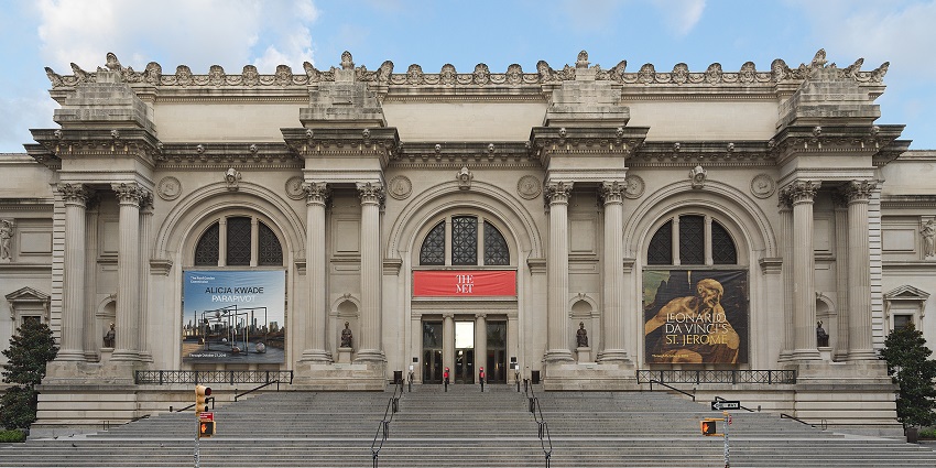 Front entrance of The Metropolitan Museum of Art in New York City with grand columns, wide steps, and visitors entering under red banners.