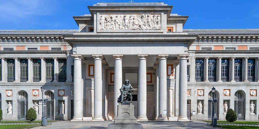 An image of Madrid's Museo del Prado entrance with classical columns, detailed pediment, flag, and statues.