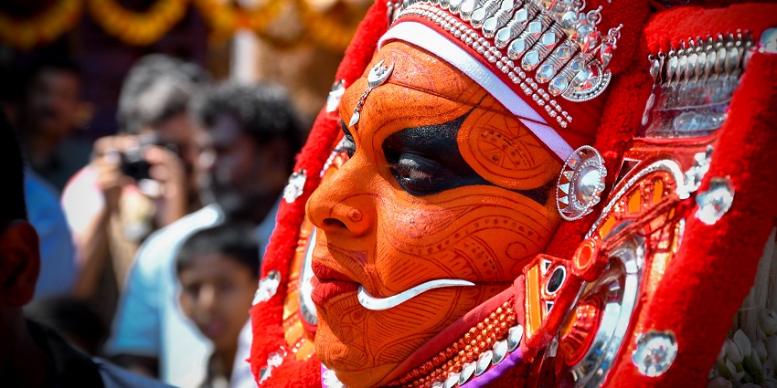 An image of a person dressed in elaborate traditional attire and jewelry, amidst a festival crowd during the Theyyam festival.
