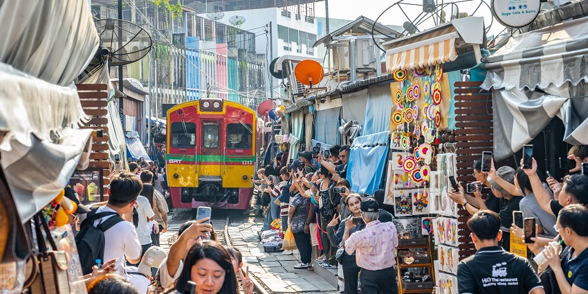 A picture of a train moving through Maeklong Railway Market, with tourists capturing the moment.