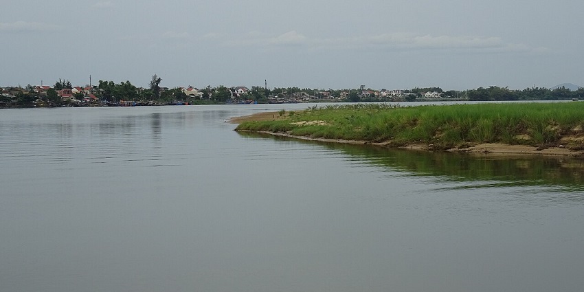 An image of the Thu Bon River in Vietnam, with calm waters reflecting the sky and surrounding greenery