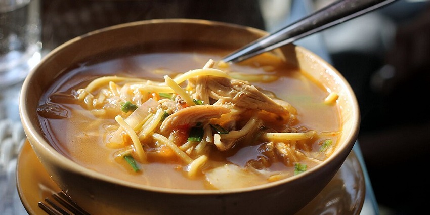 An image of a bowl of thukpa soup with noodles, vegetables, and chicken, with a spoon inside