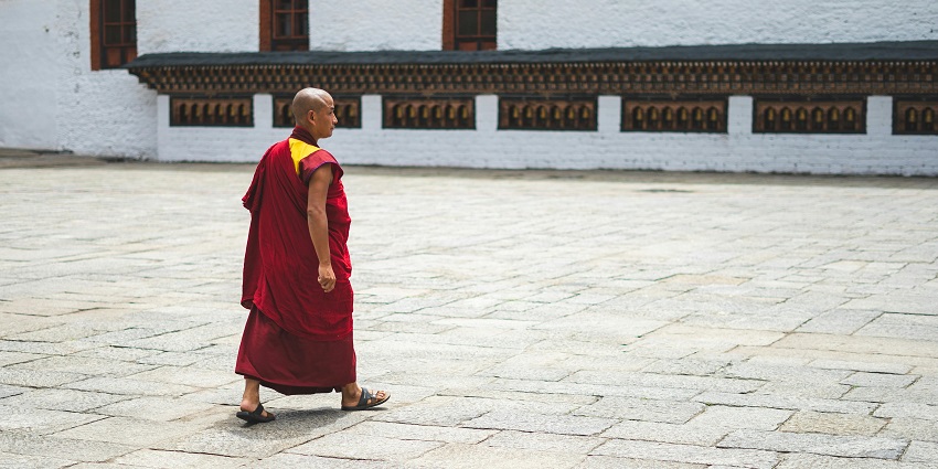An image of a monk in red and yellow robes walking a stone courtyard in a monastery resembling the Kila Gompa Nunnery